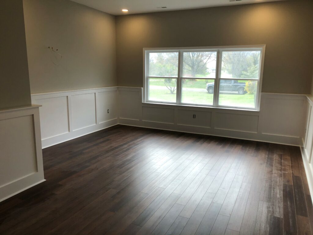 A large room with unobtrusive recessed lighting, wainscoting, a large window and gleaming hardwood floors.