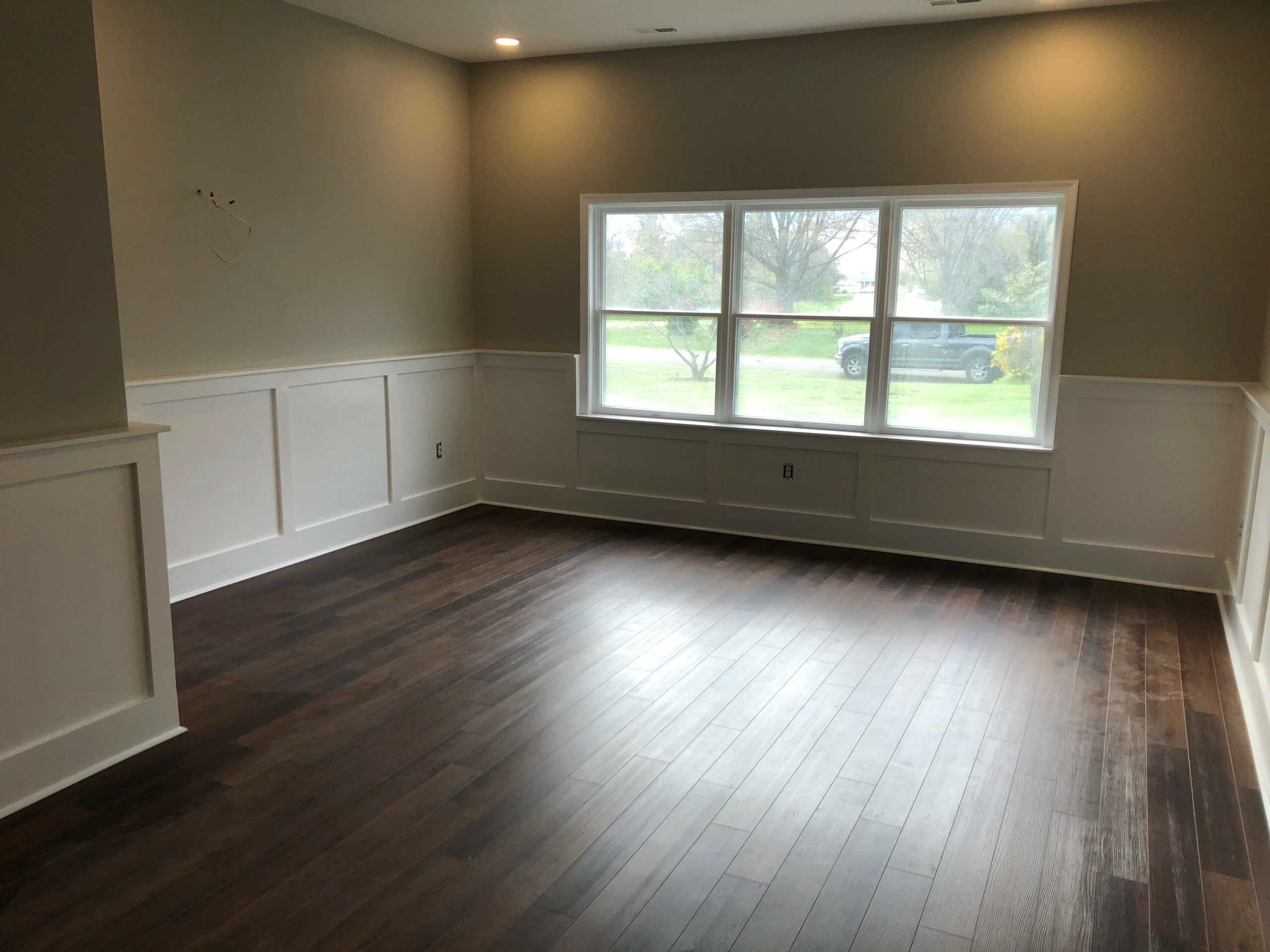 A large room with unobtrusive recessed lighting, wainscoting, a large window and gleaming hardwood floors.